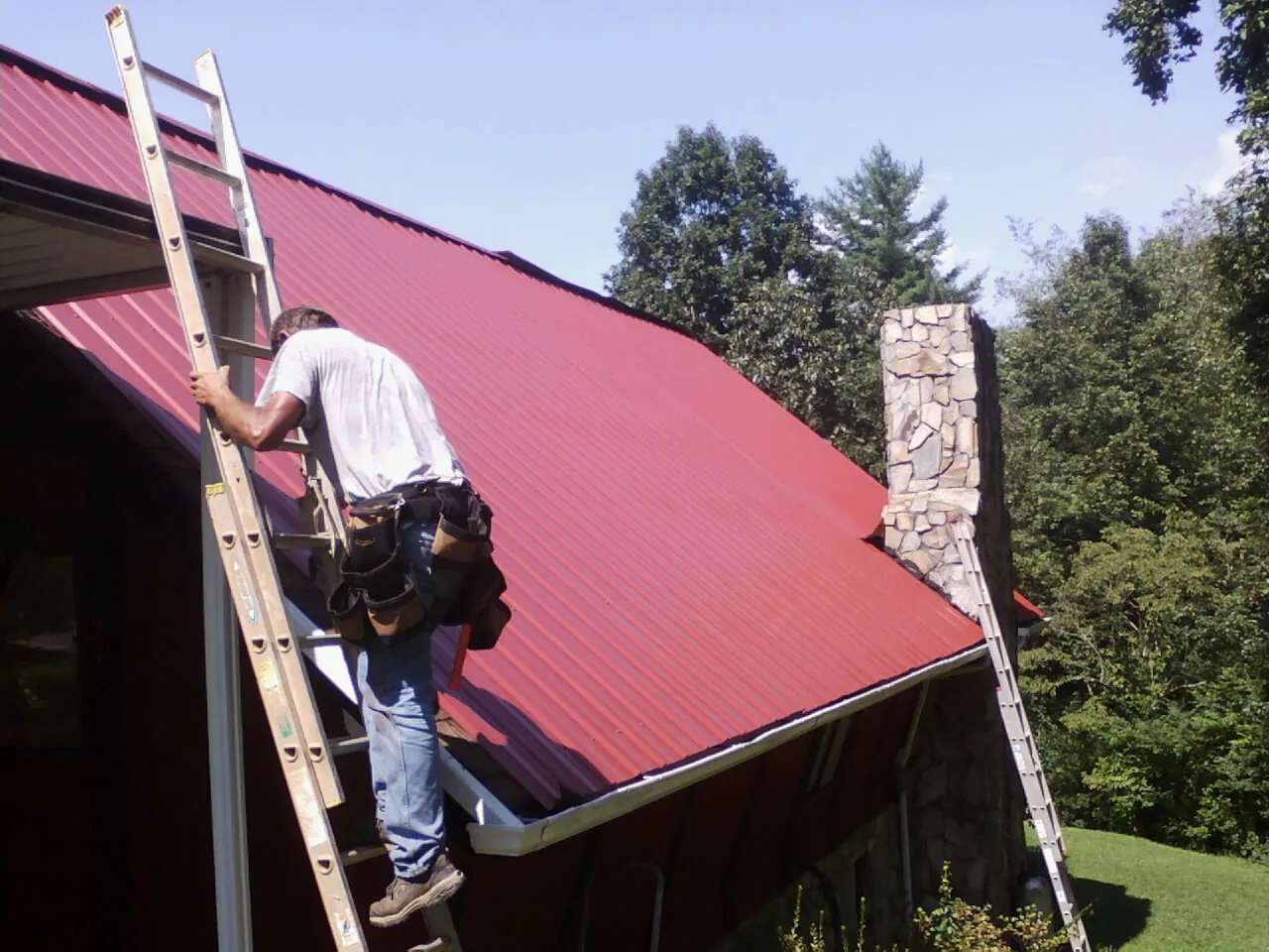 Roofer on a ladder beside a red metal roof and stone chimney — Soffit & Fascia Repair by Roofing Brothers in Whitman